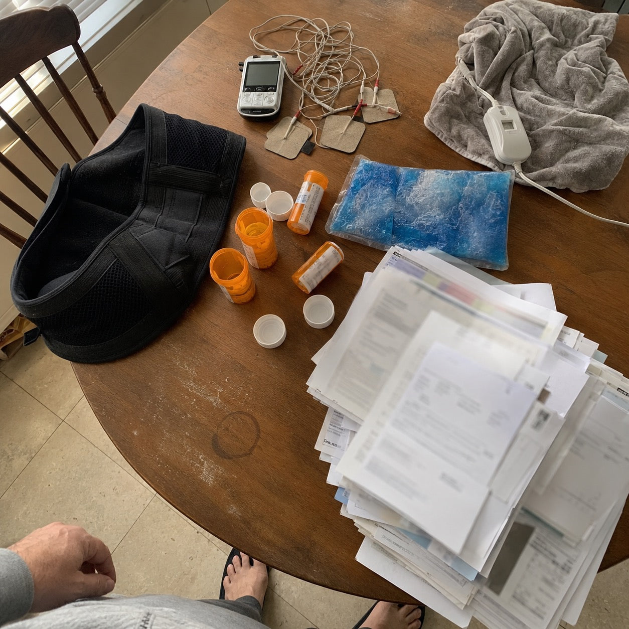 A table covered with a back brace, TENS unit, heating pad, ice pack, pill bottles and a stack of medical bills